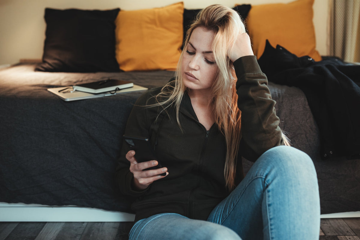 Femme assise près de son lit, regardant son téléphone d'un air pensif